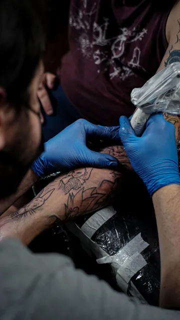 Gloved artist with a bagged machine tattooing an arm on a wrapped armrest