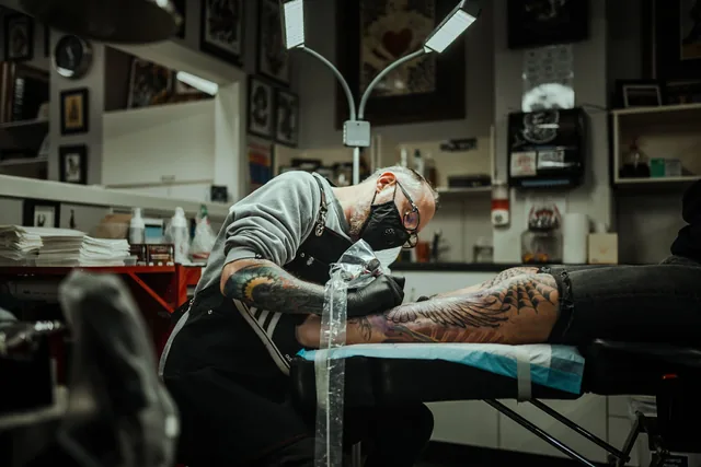 Masked, gloved artist with a bagged machine tattooing a client on a covered table