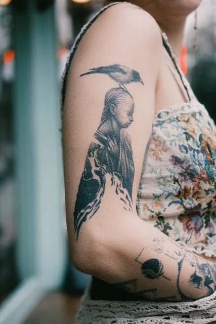 Black and gray illustrative tattoo of a figure with a crow on the upper arm, healed