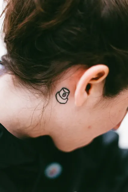 Small bold-line spray paint cap tattoo behind the ear, healed